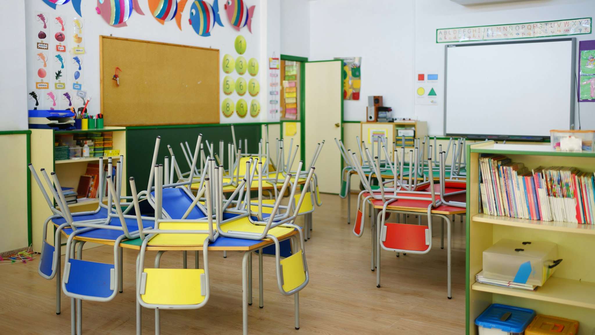 Wide view of a children's classroom at the school. Red, yellow and blue chairs arranged on the tables, white board and other school equipment.