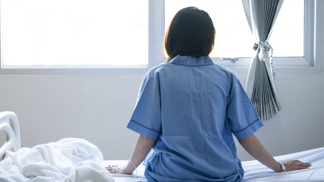 Back view of patient woman sitting on bed in hospital ward, looking away at window and hope everything will be better. -