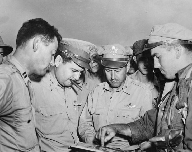 (Original Caption) With cigar in mouth, Major General Curtis E. LeMay, the commander of 21st Bomber Command, listens as B-29 Navigator Lt. Nelson McDowell (R), of Philadelphia, Pennsylvania, describes run of target after a Superfort raid on Nagoya, Japan. At left is Captain Waldo Timm, of Portland, Oregon, and second from right is Brigadier General Thomas Power, who commands B-29s on Guam.