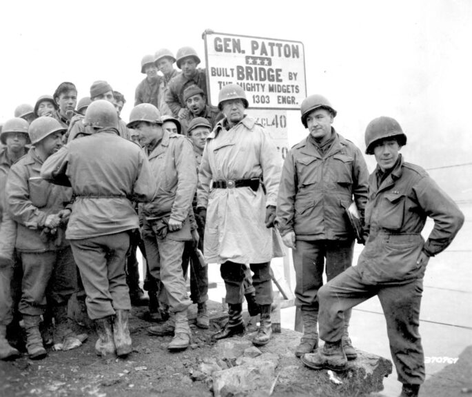 American military commander Lieutenant General George S. Patton (1885 - 1945) (center, in helmet with three stars), Commander of the 3rd Army, visits men of 1303rd Engineers, whose completed bridge across the Sauer River linked Luxembourg and Germany, February 20, 1945. The sign reads 'Gen. Patton Bridge; Built by the MIghty Midgets [obscured] 1303 Engr.' (Photo by PhotoQuest/Getty Images)