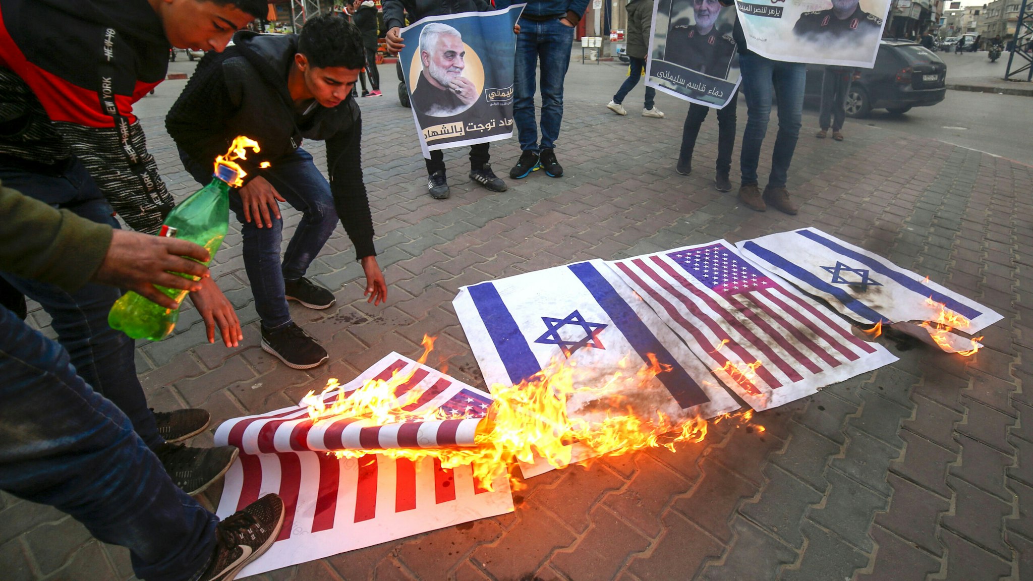 Palestinian demonstrators burn a US and an Israeli flag during a gathering in Khan Yunis in the southern Gaza Strip on January 8, 2020, organised in honour of slain Iranian military commander Qasem Soleimani (portrait) and to celebrate a volley of missiles fired by Iran at Iraqi bases housing US and other foreign troops. - Portraits of Qasem Soleimani have been carried aloft in rallies from Gaza to Yemen, raising the prospect that his violent death will elevate him as an icon of anti-American resistance.
