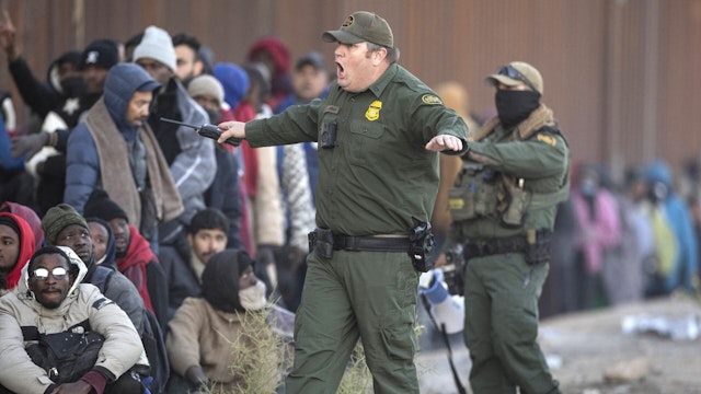 LUKEVILLE, ARIZONA - DECEMBER 06: A U.S. Border Patrol agent shouts at immigrants who had cut into a long line of people awaiting transport from the U.S.-Mexico border on December 06, 2023 in Lukeville, Arizona. A surge of immigrants illegally passing through openings cut by smugglers in the border wall has overwhelmed U.S. immigration authorities, causing them to shut down the international port of entry in Lukeville, so that officers can help process the new arrivals.