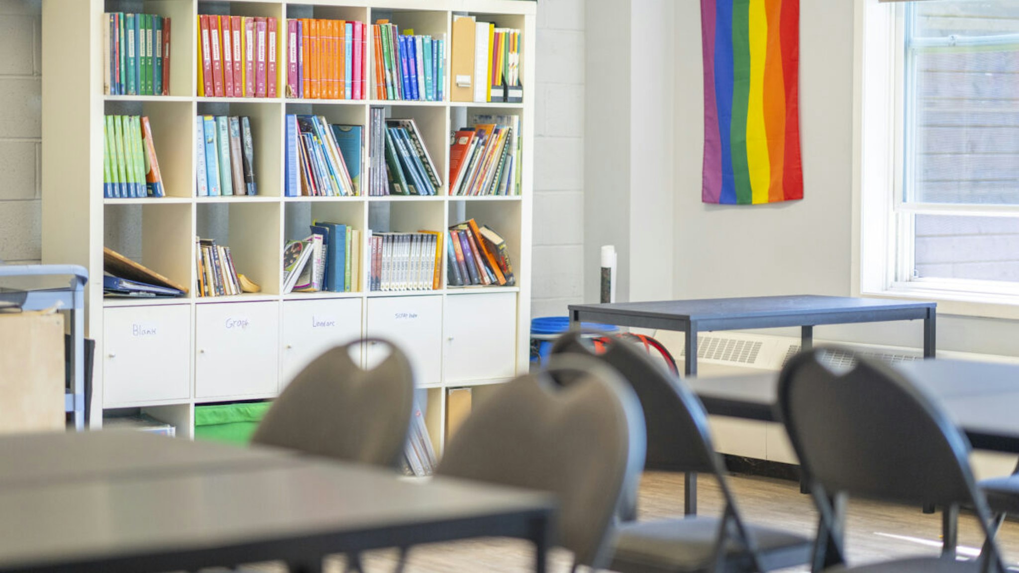 A teachers corner of a Montessori classroom with bookshelves containing binders and textbooks, a small desk and a pride flag on the wall. Empty student desks can be seen in the foreground.