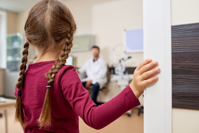 Back view portrait of scared little girl entering doctors office, standing in doorway and looking at blurred pediatrician