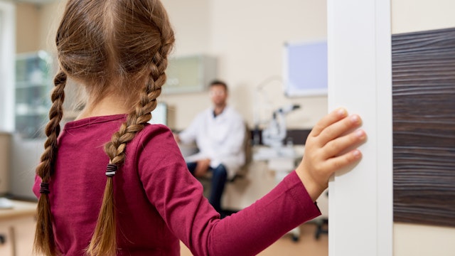 Back view portrait of scared little girl entering doctors office, standing in doorway and looking at blurred pediatrician