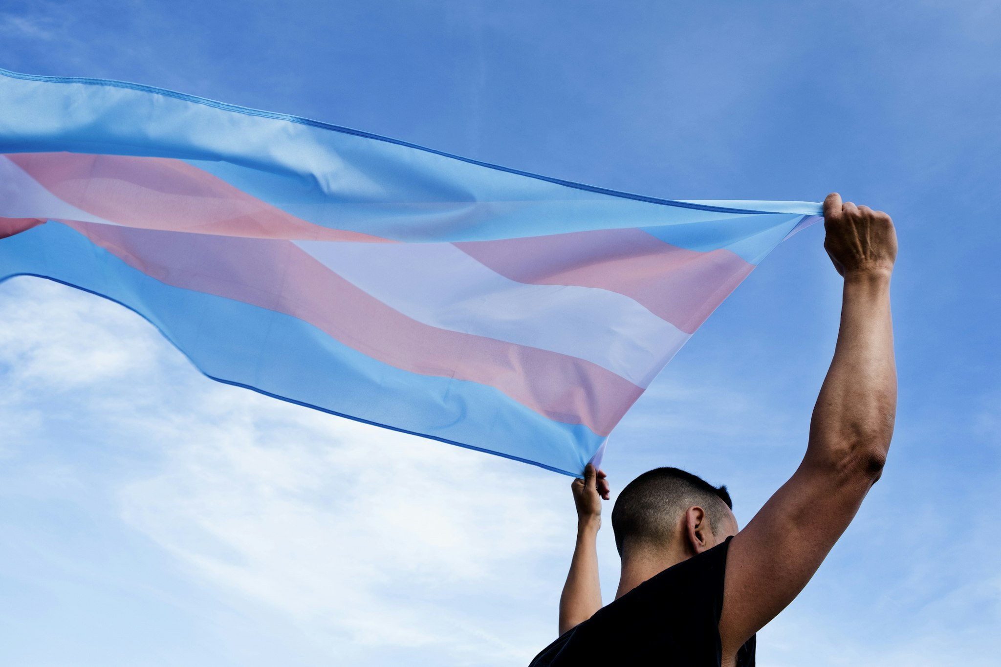 nito100. Getty Images. a young caucasian person, seen from behind, holding a transgender pride flag over his or her head against the blue sky