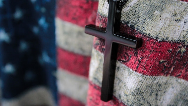 Wirestock. Getty Images. A small wooden cross and flag of USA. God save America.