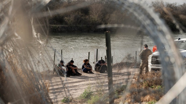 Migrants wait with members of the Texas National Guard to be processed by border patrol along the banks of the Rio Grande River in Eagle Pass, Texas, US, on Friday, Feb. 23, 2024. The US Border Patrol will be forced to make operational cuts if lawmakers don't authorize funding to address the border crisis, the agency's chief has warned.