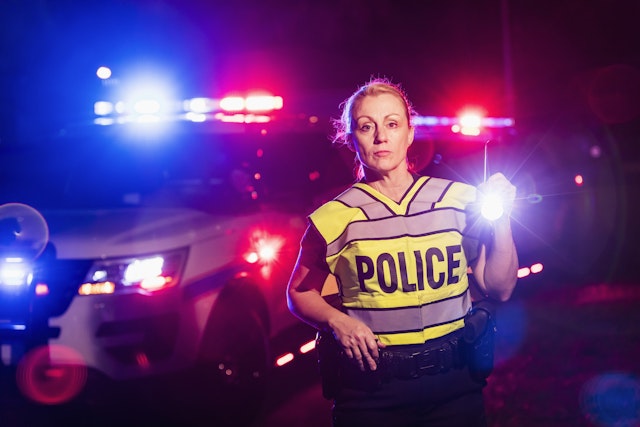 kali9. Getty Images. A policewoman wearing a reflective vest, standing in front of police cars with emergency lights flashing, holding a flashlight. She is a mature woman in her 40s.