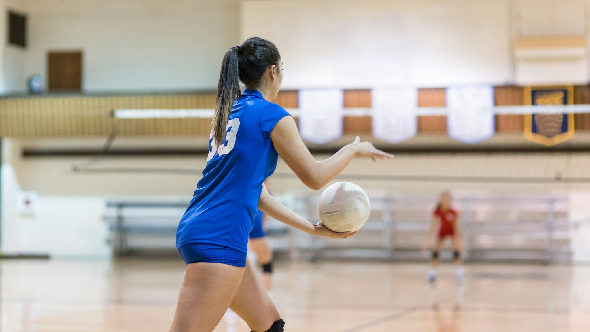 A teenage volleyball player gets ready to serve the ball in a high school gym. The shot is from behind her. The opposing team is getting ready to return the serve. There are banners hanging in the gym overhead.