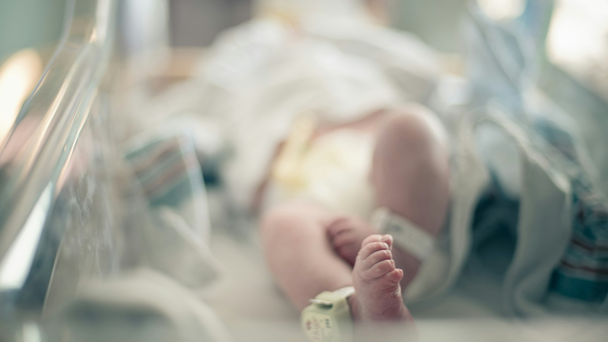 Newborn baby lies on its back surrounded by blankets in hospital bassinet with only its feet and ankle tracking bracelet in focus, photographed with shallow depth of field allowing for ad space.