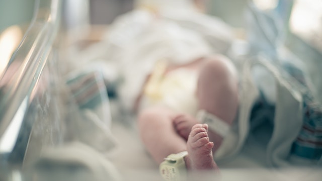 Newborn baby lies on its back surrounded by blankets in hospital bassinet with only its feet and ankle tracking bracelet in focus, photographed with shallow depth of field allowing for ad space.