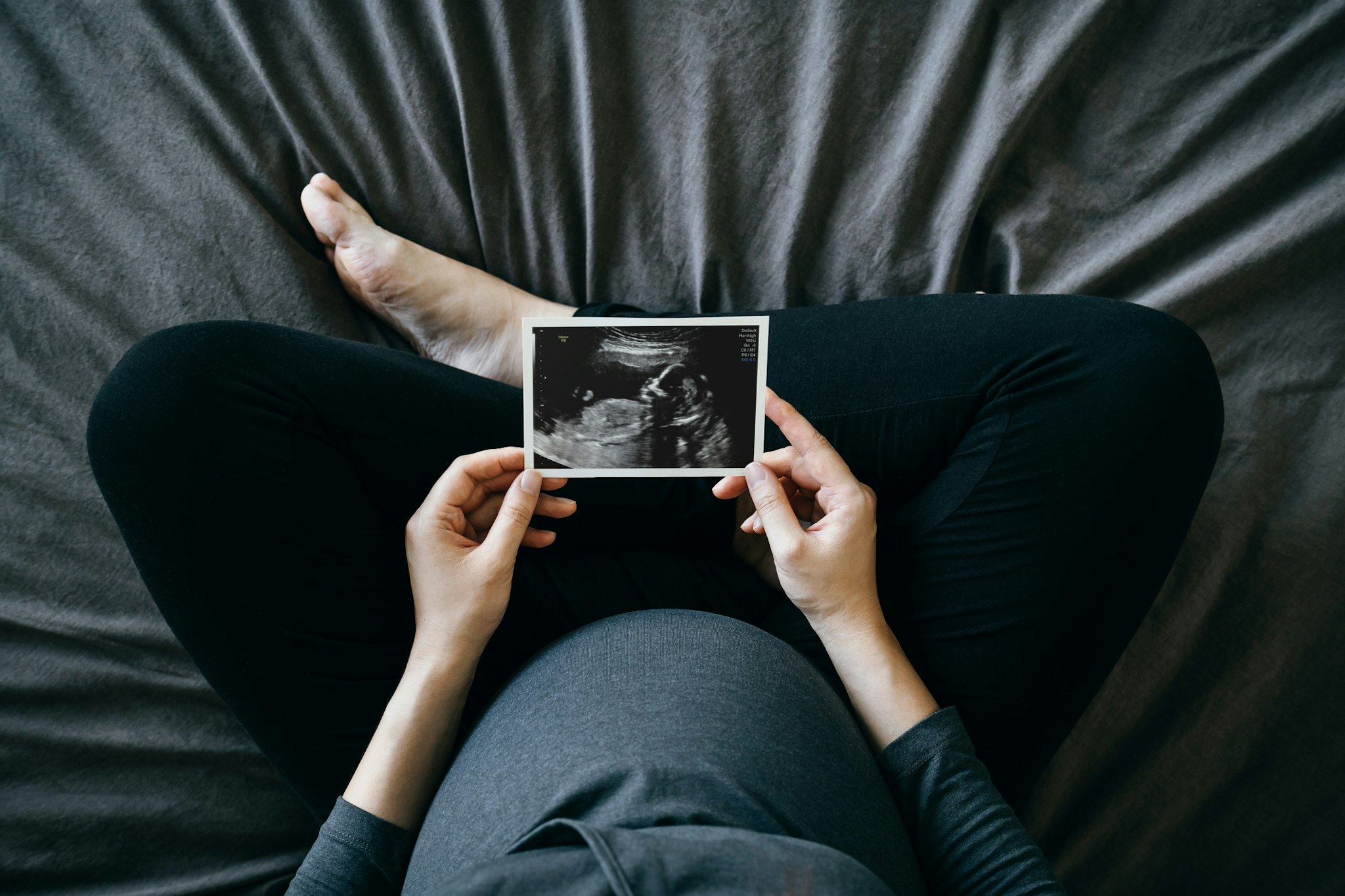 d3sign. Getty Images. High angle shot of Asian pregnant woman holding an ultrasound scan photo in front of her baby bump, sitting on bed at home. Mother-to-be. Precious moment in life. Preparation for a new family member. Expecting a new life. Baby and new life concept