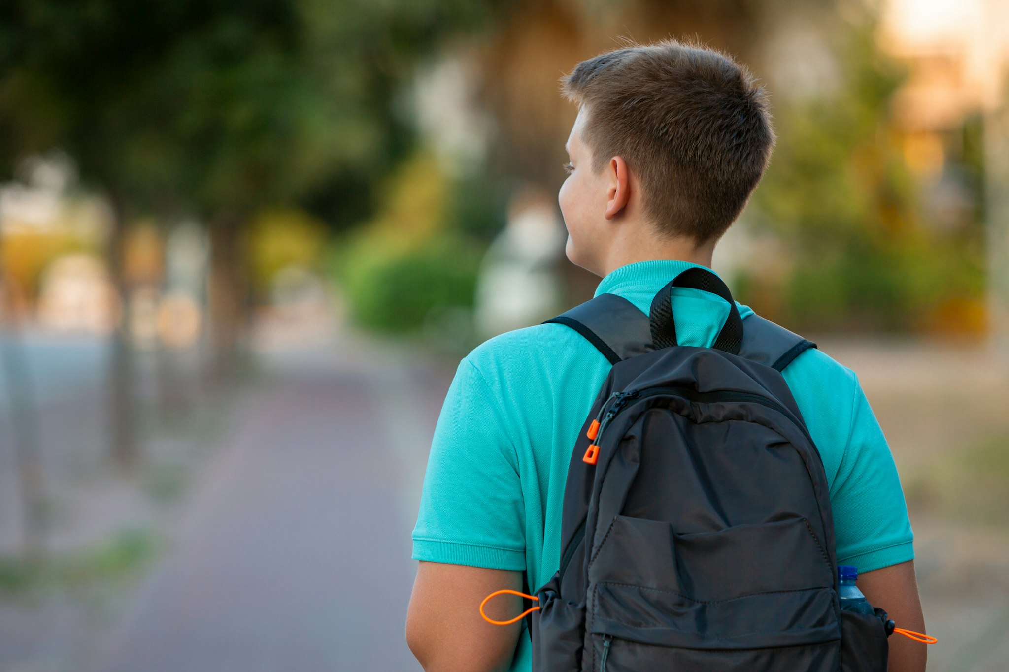 Radiating enthusiasm and dressed in a vibrant turquoise T-shirt, a cheerful teen boy stands ready for school with a backpack in hand. His joyful expression reflects a genuine love for learning as he embraces the excitement of a new day. The warm glow of late daytime enhances the scene, casting a positive light on this junior adventurer's journey into the world of education. This image captures the spirit of youthful exuberance and the anticipation of new discoveries in the school environment.