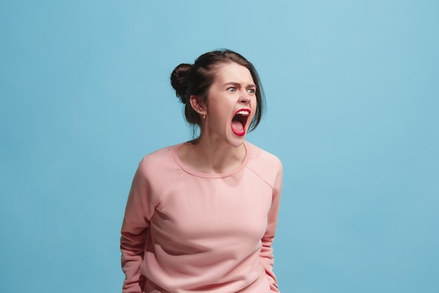 master1305. Getty Images. Screaming, hate, rage. Crying emotional angry woman screaming on blue studio background. Emotional, young face. Female half-length portrait. Human emotions, facial expression concept. Trendy colors.