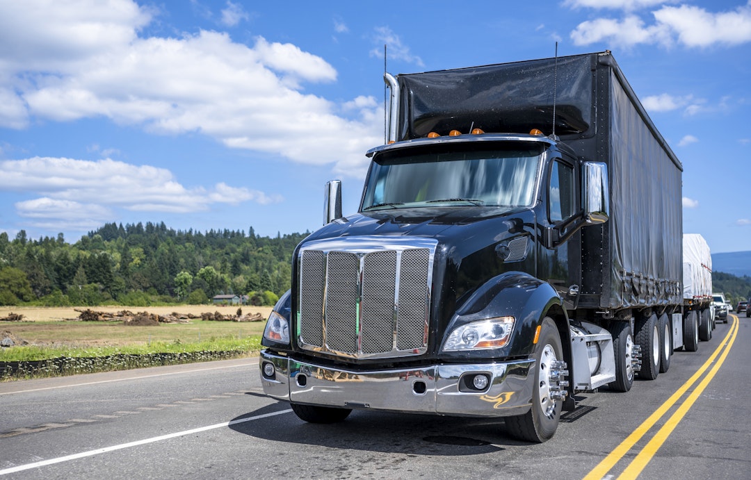 Industrial shiny black day cab professional big rig semi truck tractor transporting commercial cargo in covered dry van and on flat bed semi trailers driving on the narrow road at sunny day.