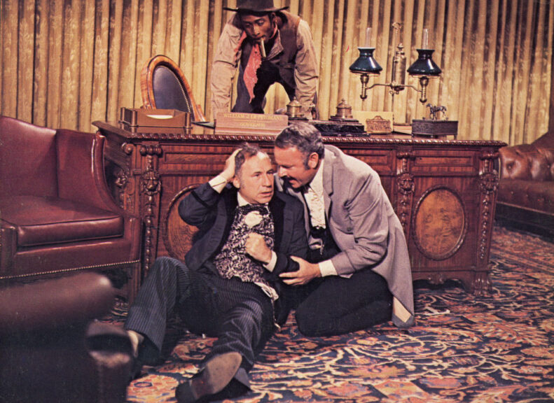 Actor Mel Brooks (left) sits on the floor beside Harvey Korman as Cleavon Little kneels atop a desk, in a still from the film, 'Blazing Saddles,' directed by Mel Brooks, 1974. (Photo by Warner Bros./Courtesy of Getty Images)