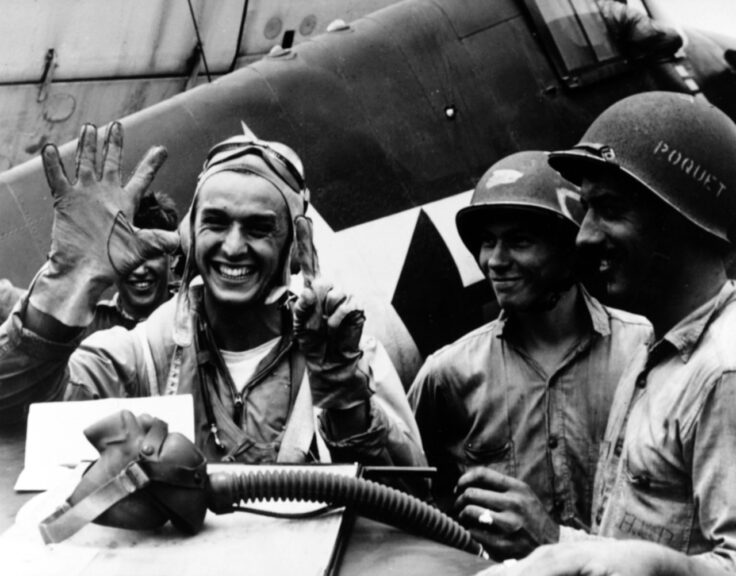 Lieutenant Junior Grade Alexander Vraciu, USNR; fighting squadron 16 "Ace", holds up six fingers to signify his "kills" during the "Great Marianas Turkey Shoot", on 19 June 1944. Taken on the flight deck of the USS LEXINGTON (CV-16). Note: Grumman in background and sailor A.L. Poquet at right. Catalog #: 80-G-236841. National Archives. Original Date: Mon, Jun 19, 1944. Naval History and Heritage Command.