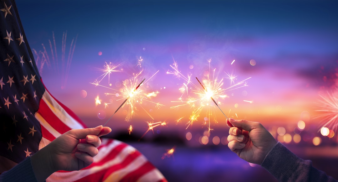 RomoloTavani. Getty Images. Usa Celebration With Hands Holding Sparklers And American Flag At Sunset With Fireworks.