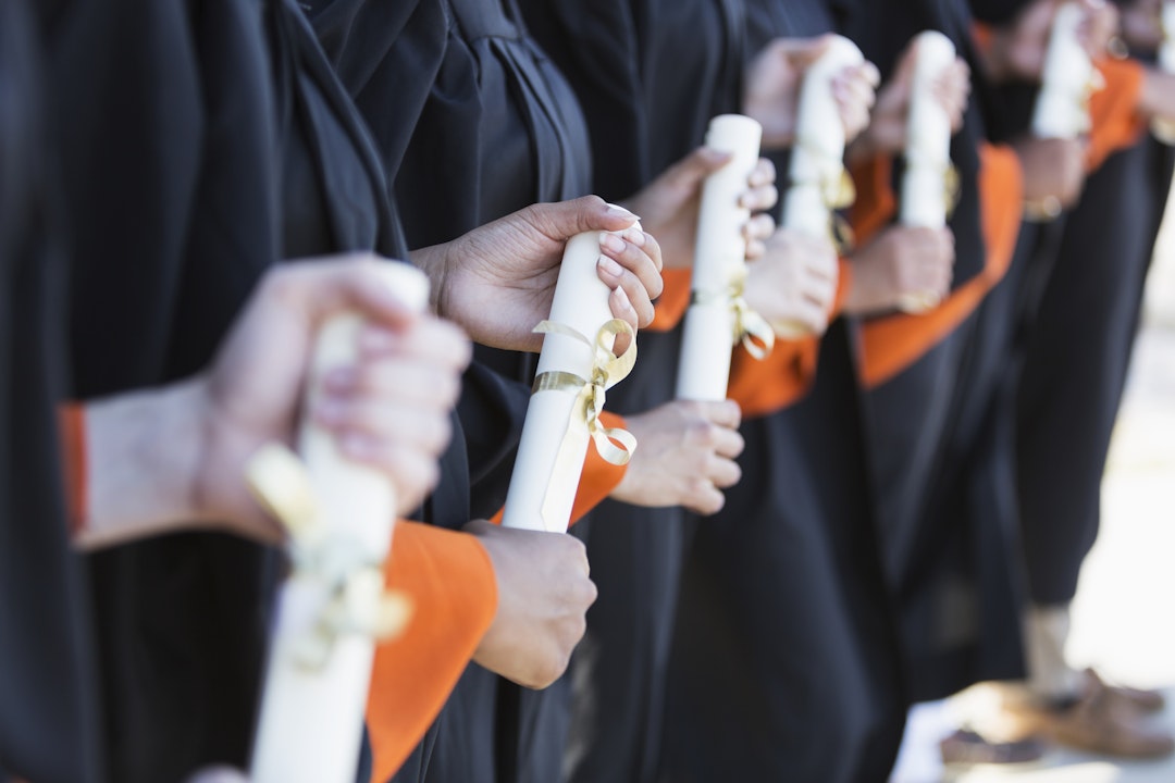 kali9. Getty Images. Cropped view of a group of multi-ethnic students graduating from high school or university. They are standing outdoors in a row, wearing black graduation gowns, holding diplomas.