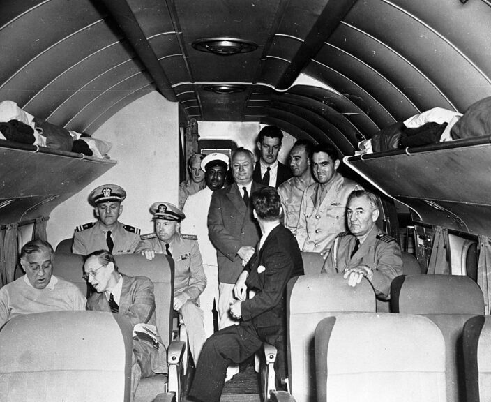 Homeward bound from the Casablanca Conference of 1943, President Roosevelt is shown in animated conference with Harry Hopkins. Sitting directly behind them are Lieutenant George Fox (left), US Navy Medical Corps, and Rear Admiral Ross T. McIntyre (right), Surgeon General of Navy. Across the aisle are Guy H. Spaman (back to camera) of the US Secret Service, and Captain John L. McCrea, Presidential Naval Aide. Standing are Arthur Prettyman, valet, Charles Fredericks, (hands folded), Elmer Hipley, Secret Service, Wilmer Deekand, Secret Service, and Captain Otis Bryan, Chief Pilot.